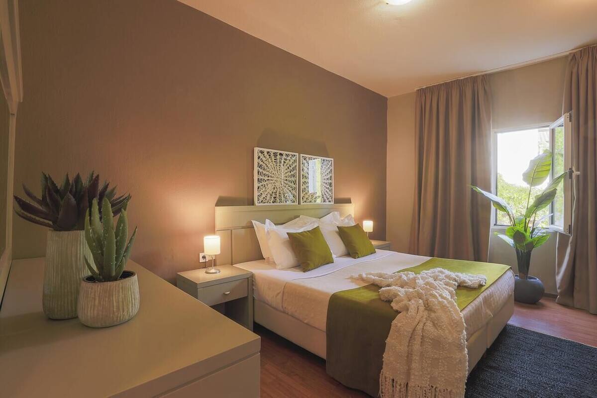 A bedroom with a bed, bedside tables, a nightstand, a decorative wall piece, and potted plants.