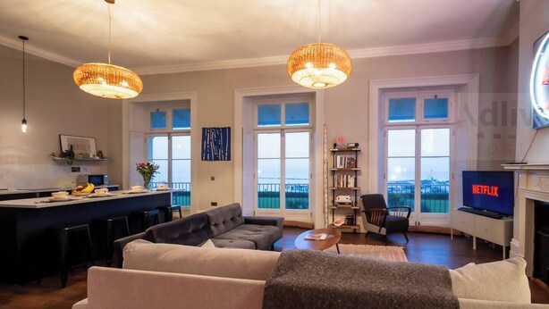A modern living room with a sofa, a coffee table, and a TV displaying 'NETFLIX'. There are two large windows offering a view of the city. - Historic Seaview Gem in Ramsgate by Adliv (RAMSGATE)