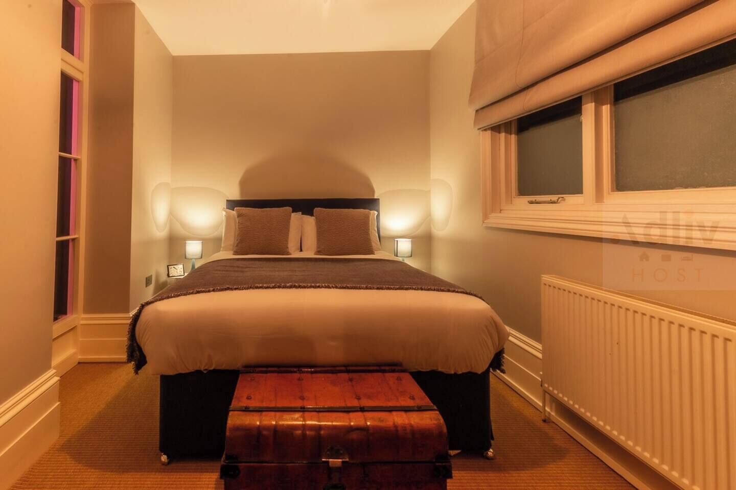 A bedroom with a bed, bedside lamps, a window, a radiator, and a wooden bench.