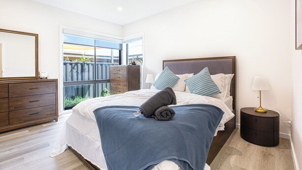 A bedroom with a bed, a dresser, a bedside table with a lamp, and a window with a view of greenery. - Modern 3B house, pet friendly close nature reserve (Throsby)