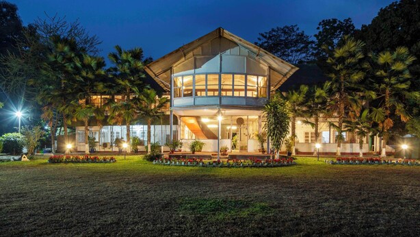 A two-story building with a glass facade, surrounded by a well-maintained garden with red flowers and palm trees. - amã Stays & Trails Hathikuli, Assam (Bokakhat)