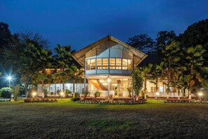 A two-story building with a glass facade, surrounded by a well-maintained garden with red flowers and palm trees. - amã Stays & Trails Hathikuli, Assam (Bokakhat)