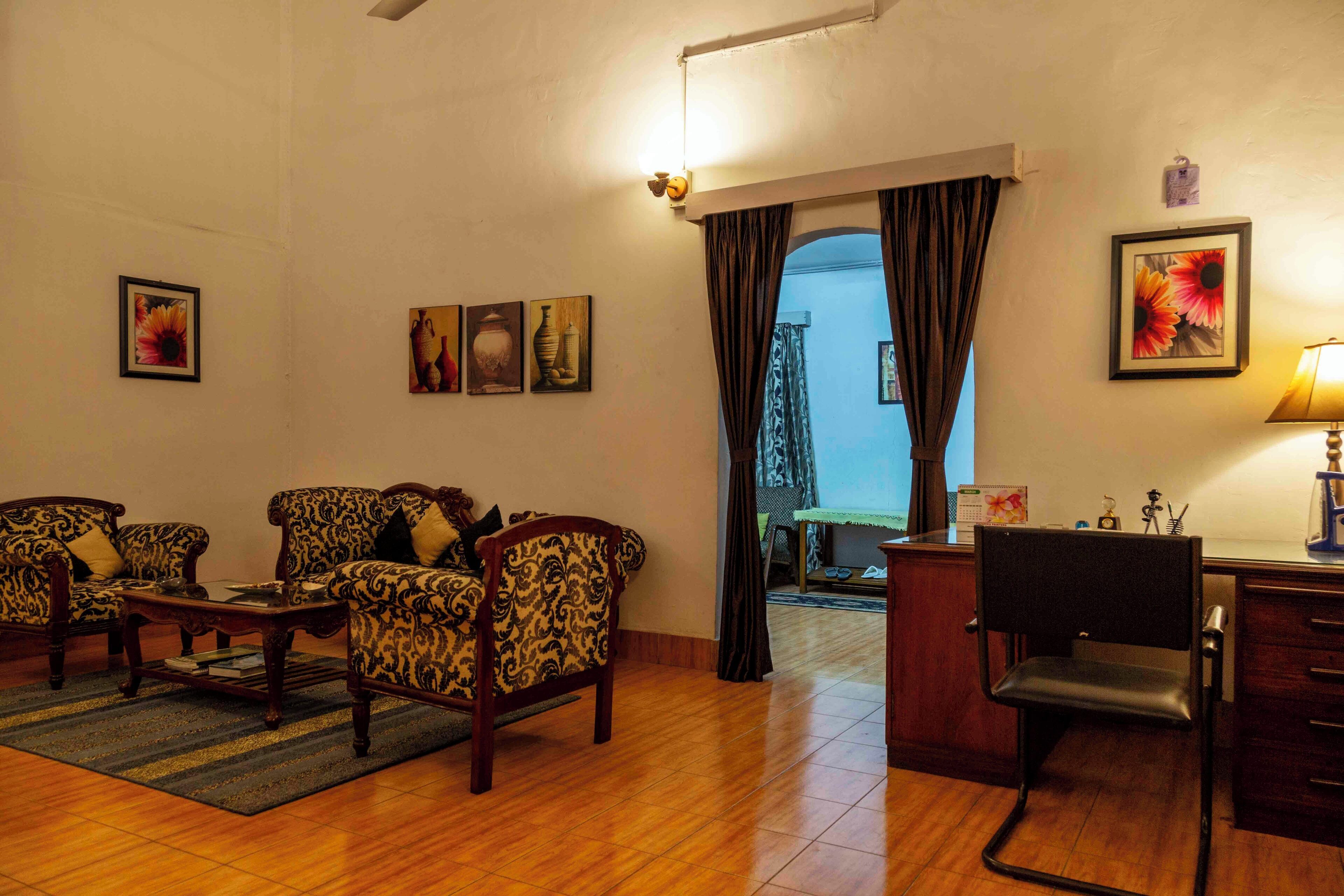 A living room with patterned sofas, a wooden coffee table, a desk with a chair, and a lamp. There are framed pictures on the wall and a view through a doorway.