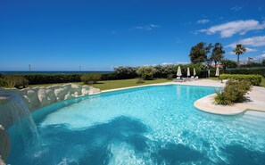 Une piscine d’un bleu éclatant, bordée de chaises longues et d’ombrelles, entourée de verdure et offrant une vue sur l’océan.