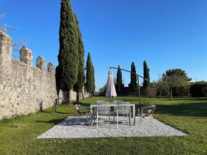 Un espacio con mesas y sillas al aire libre, rodeado por un muro de piedra y altos árboles.