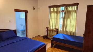 A room with two wooden beds, a wooden chair, a window with curtains, and a door leading to a bathroom.