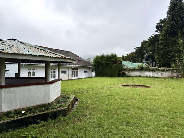 A building with a corrugated metal roof, white walls, and a small garden area with grass and a circular flower bed. - Cottage 88 (Nuwara Eliya)