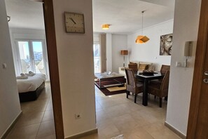 Habitación de hotel con cama, zona de comedor con mesa y sillas, un reloj en la pared y un cuadro.