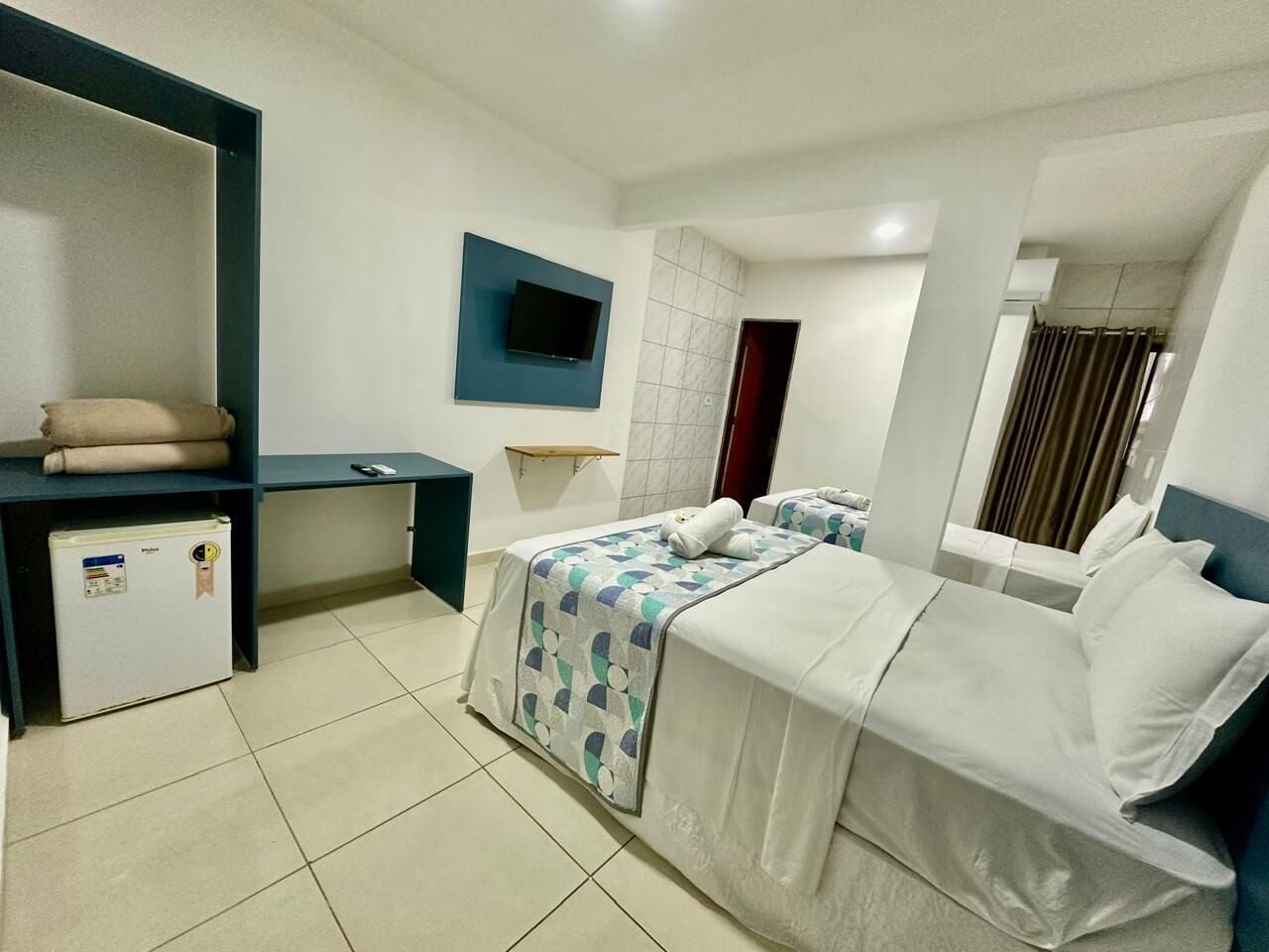 A hotel room with two beds, a desk, a TV, a mini-fridge, and a window with curtains.