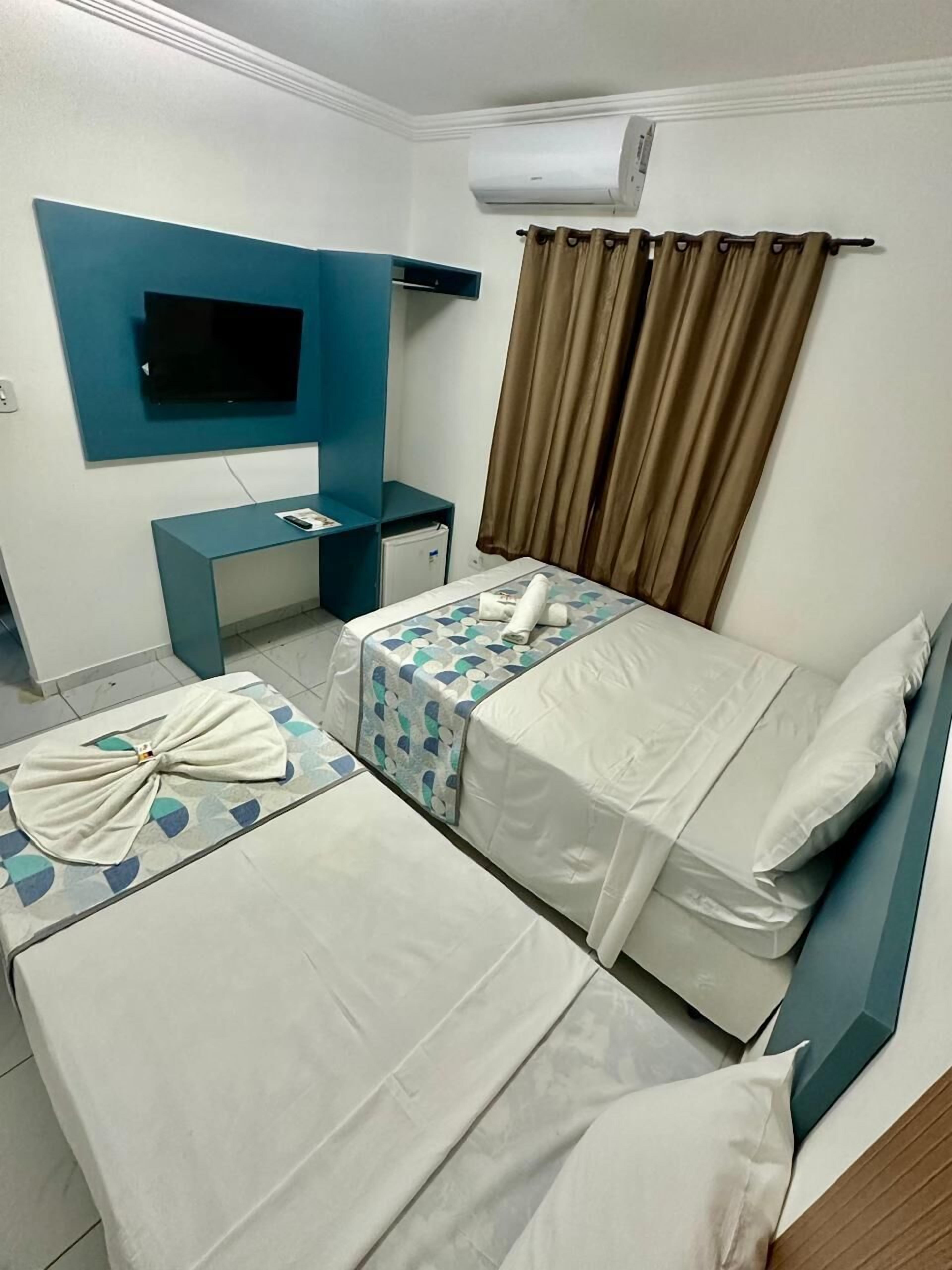 A hotel room with two single beds, a desk, a flat-screen TV mounted on the wall, and a window with curtains.