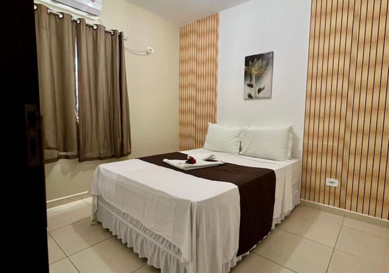 A hotel room with a bed, curtains, a wall-mounted air conditioner, and a wooden panel wall.