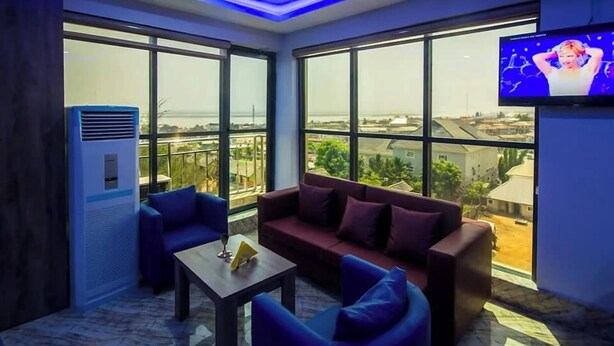 A modern room with a large window, a flat-screen TV, and a sofa. - Golden Grand Royale Hotel  (Lokoja)