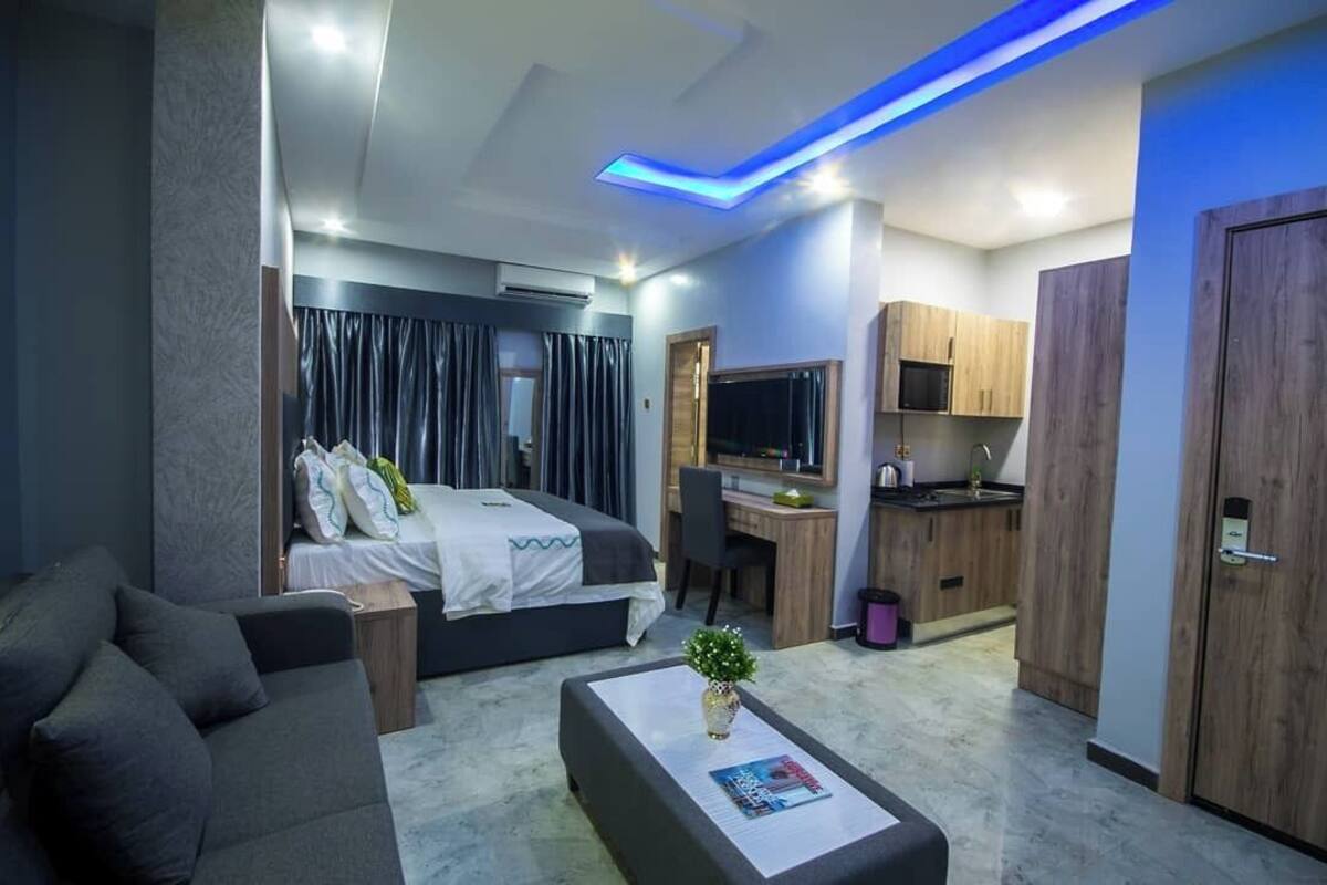 A modern hotel room with a bed, a desk, a TV, and a kitchenette.