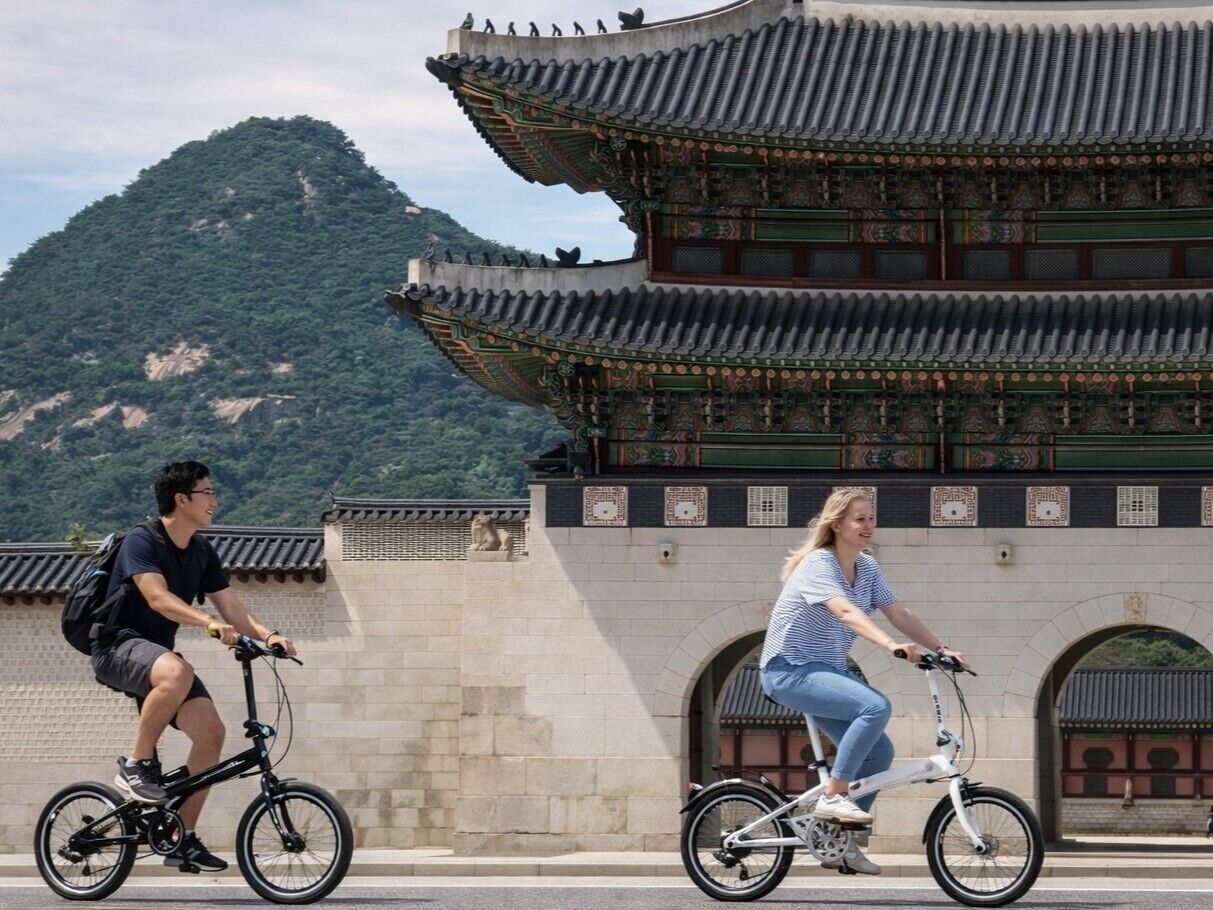 Dos personas montando bicicletas frente a un edificio tradicional con techo de tejas y paredes de piedra.
