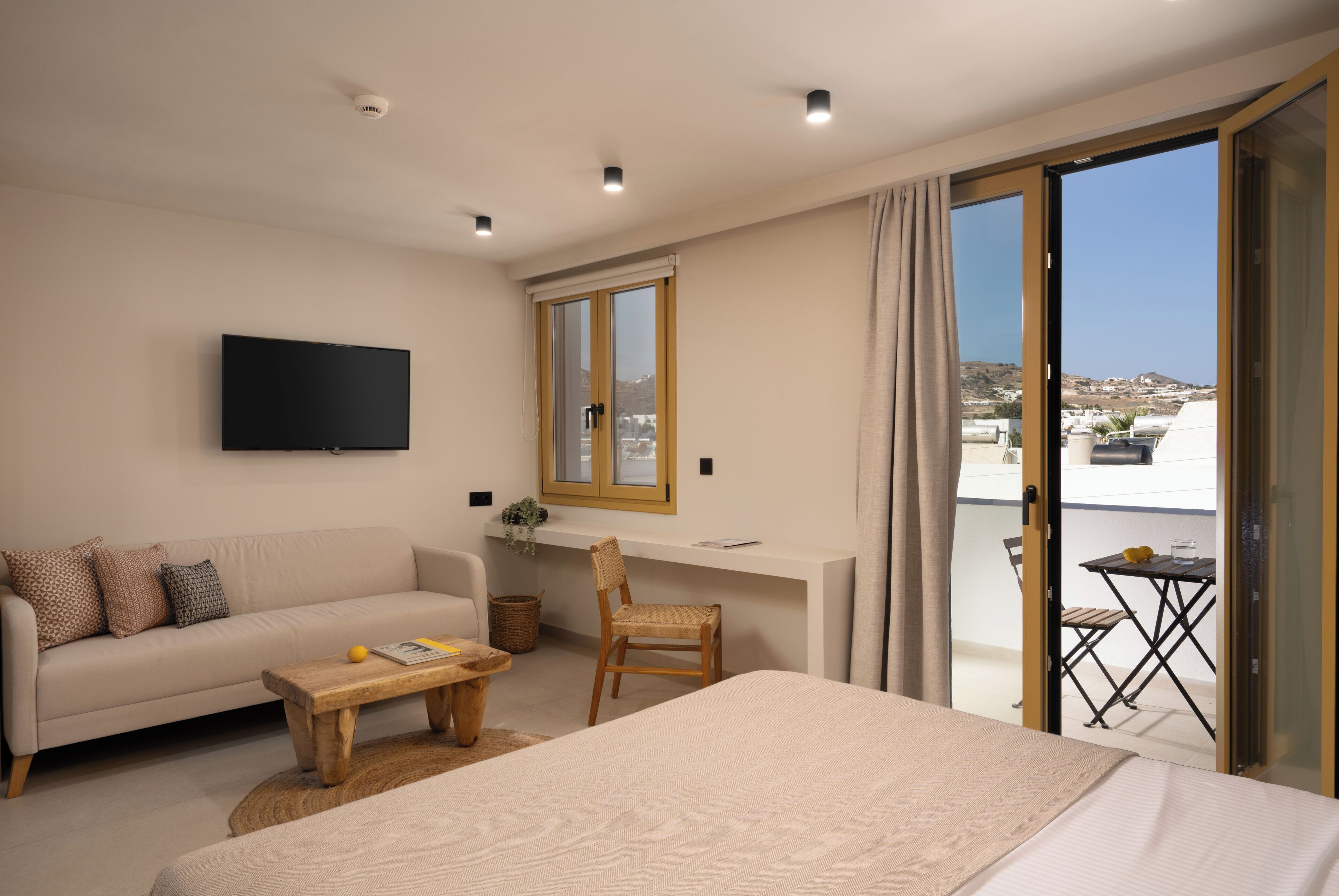 Une chambre d’hôtel moderne comprenant un lit, un canapé, une petite table, une chaise, un téléviseur fixé au mur, une fenêtre donnant sur la ville et un balcon avec une table et des chaises.