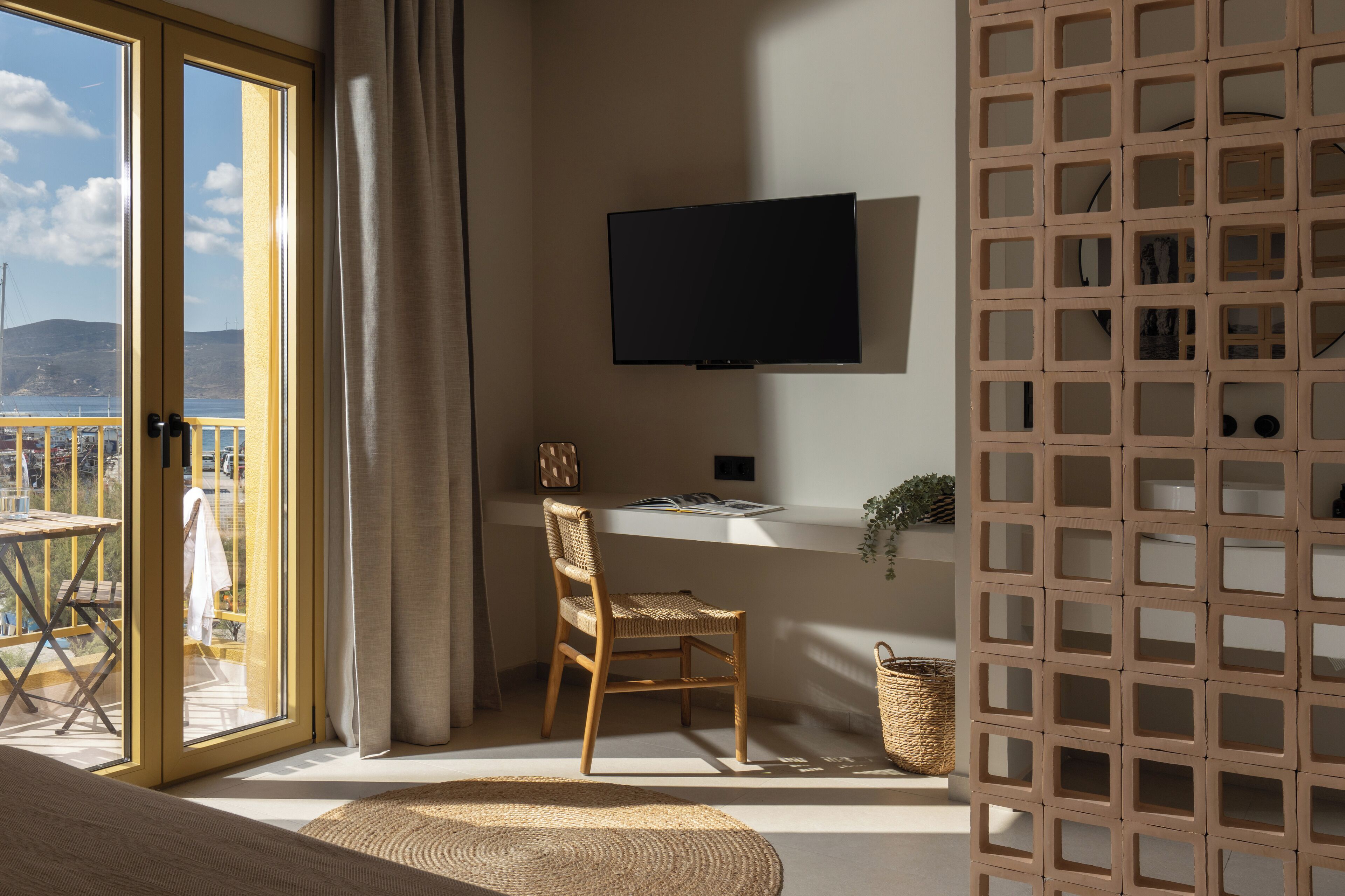 Una habitación con puerta de vidrio corredera que da a un balcón, un escritorio con silla, un televisor de pantalla plana y una cesta tejida.
