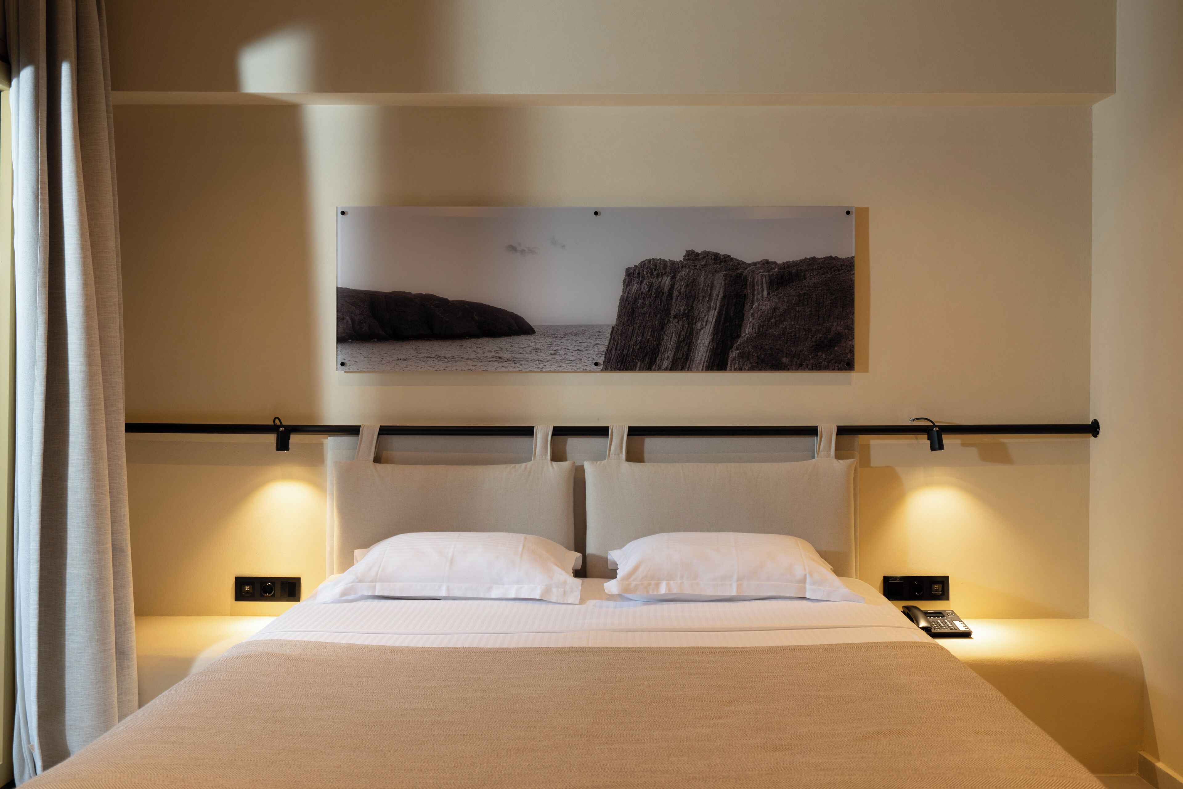 Habitación de hotel con cama, dos mesitas de noche, un cuadro colgado de un paisaje costero y una cortina a la izquierda.