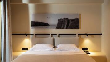 Une chambre d’hôtel avec un lit, deux tables de chevet, un tableau représentant un paysage côtier accroché au mur et un rideau sur le côté gauche.