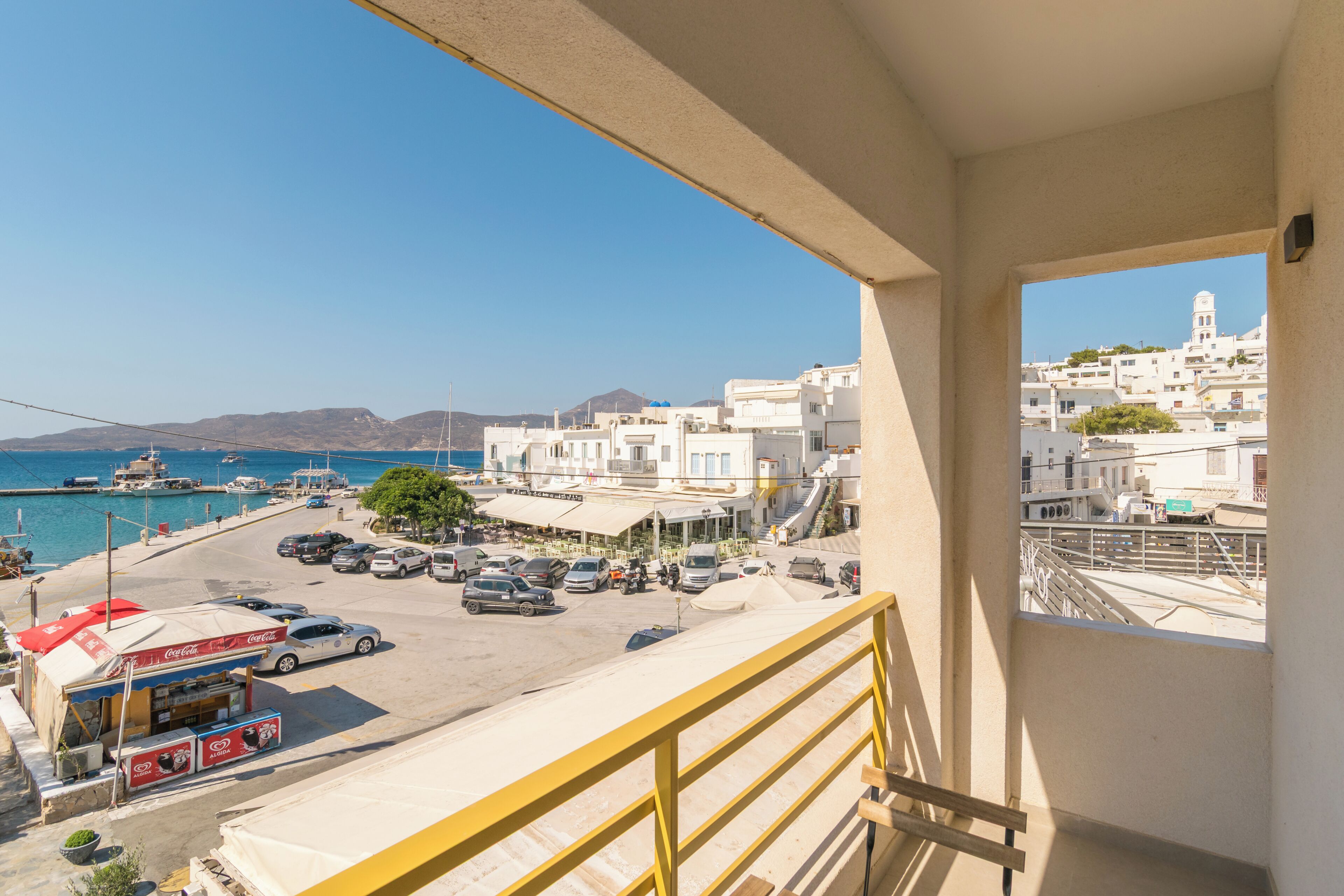 Un balcón con vista a un pueblo costero, un puerto deportivo con barcos y coches estacionados.
