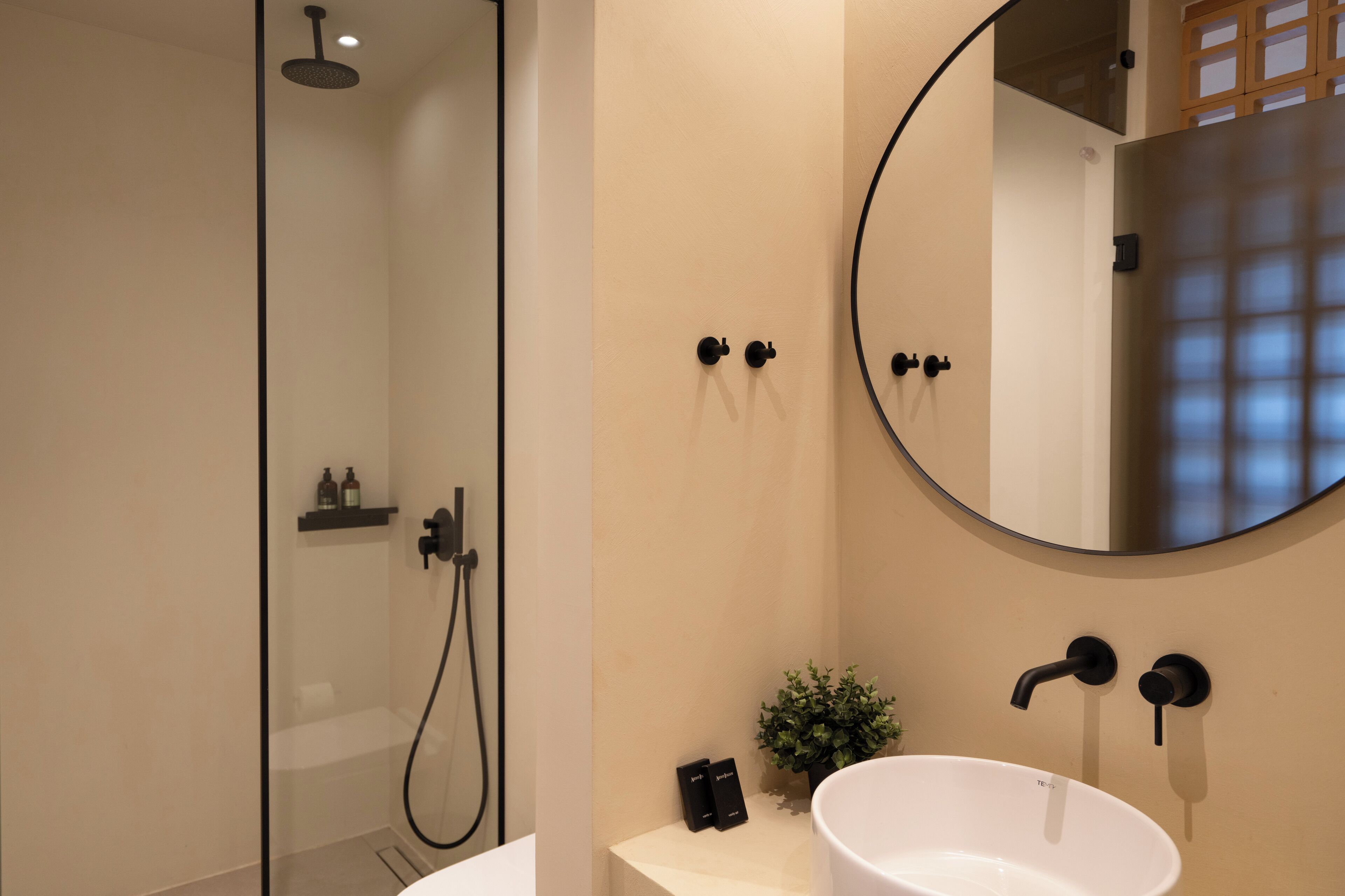 Un baño moderno con ducha de vidrio, un espejo redondo, un lavamanos blanco y una pequeña planta en maceta.