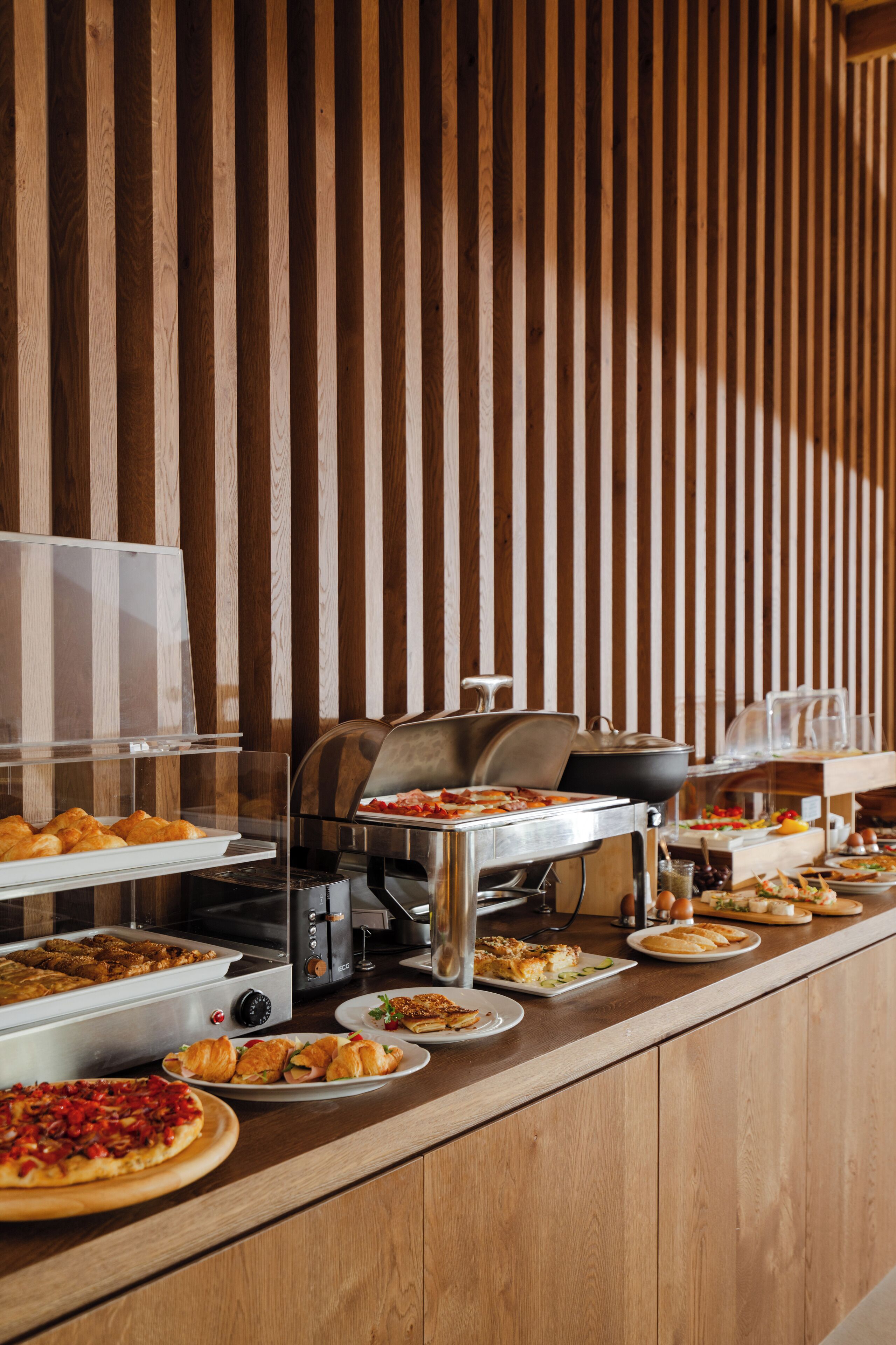 Ein Buffet mit verschiedenen Speisen, darunter Pizza, Sandwiches und Gebäck, vor rustikalem Holzhintergrund.