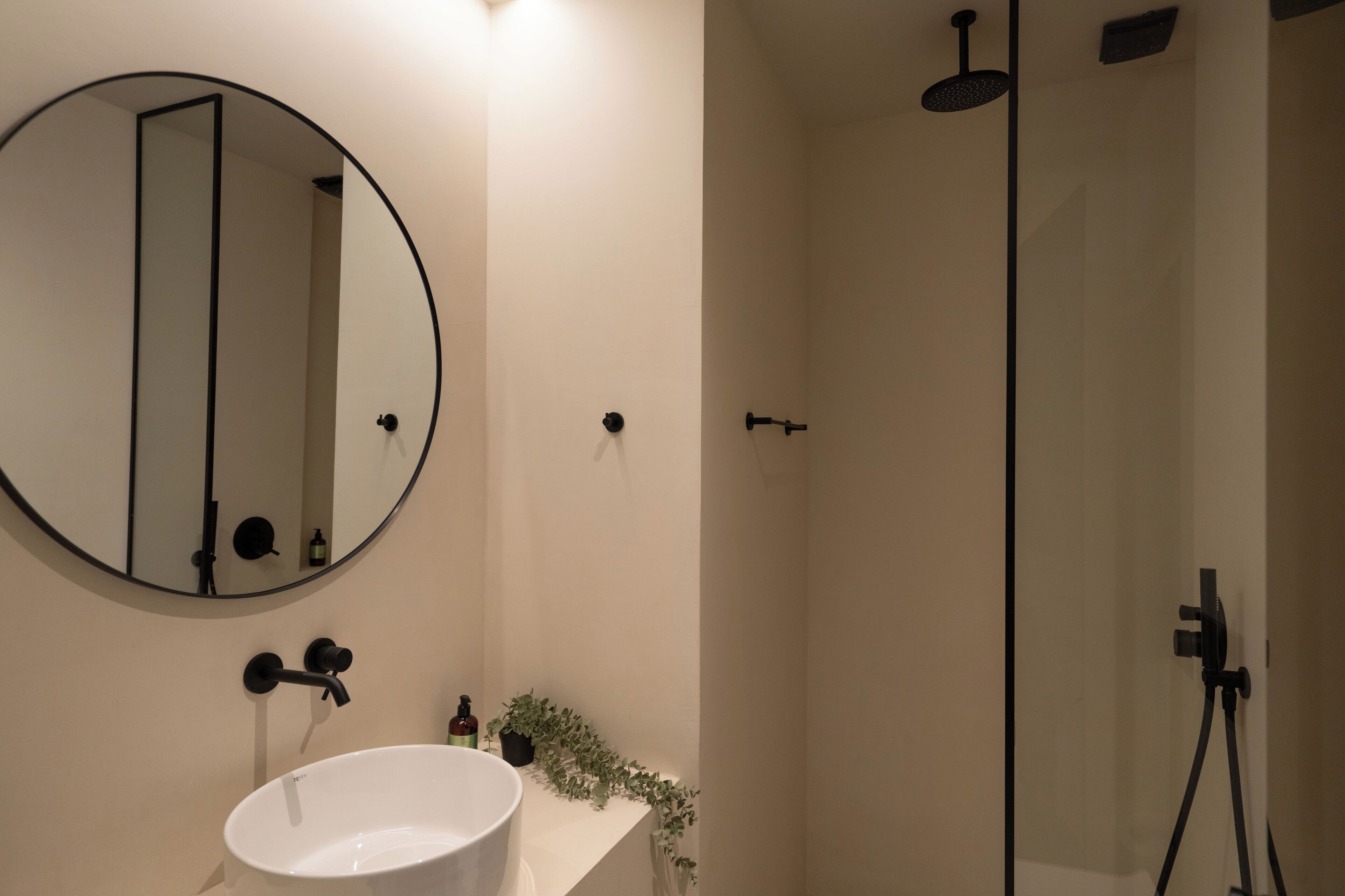 Un baño moderno con un espejo redondo, un lavabo blanco, un cabezal de ducha de mano y una pequeña planta en maceta.