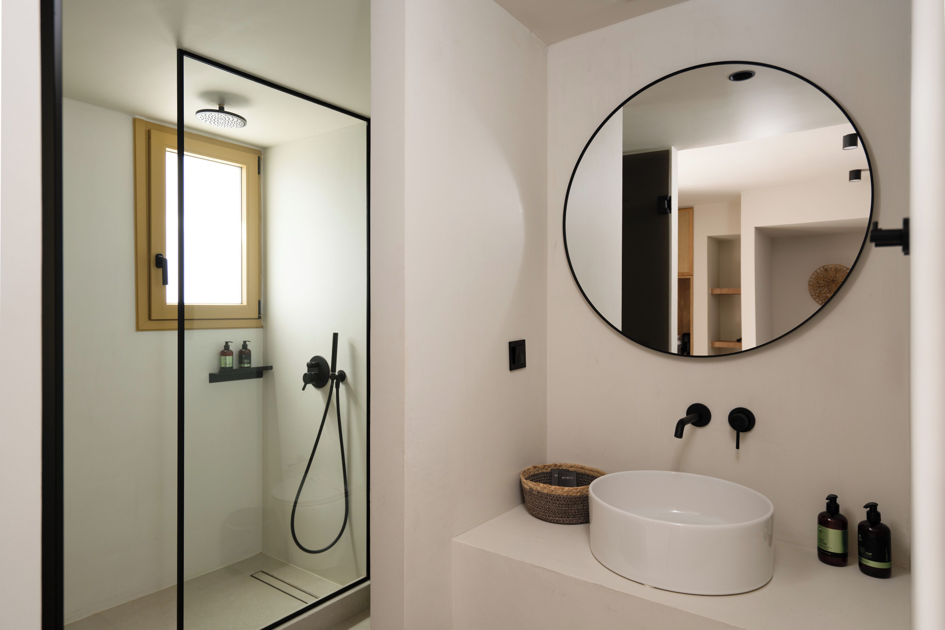 A modern bathroom with a glass-enclosed shower, a round mirror, and a white sink with two bottles of soap.