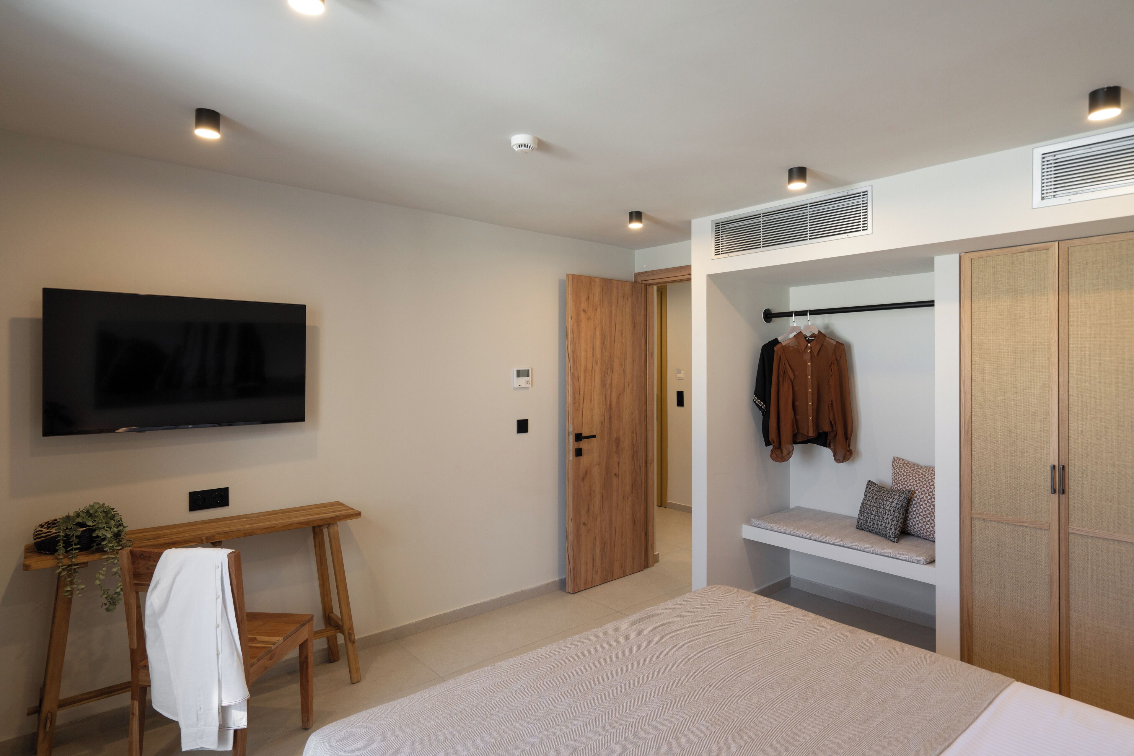 Un dormitorio moderno con una cama grande, un televisor montado en la pared, un escritorio de madera y un armario con un banco.