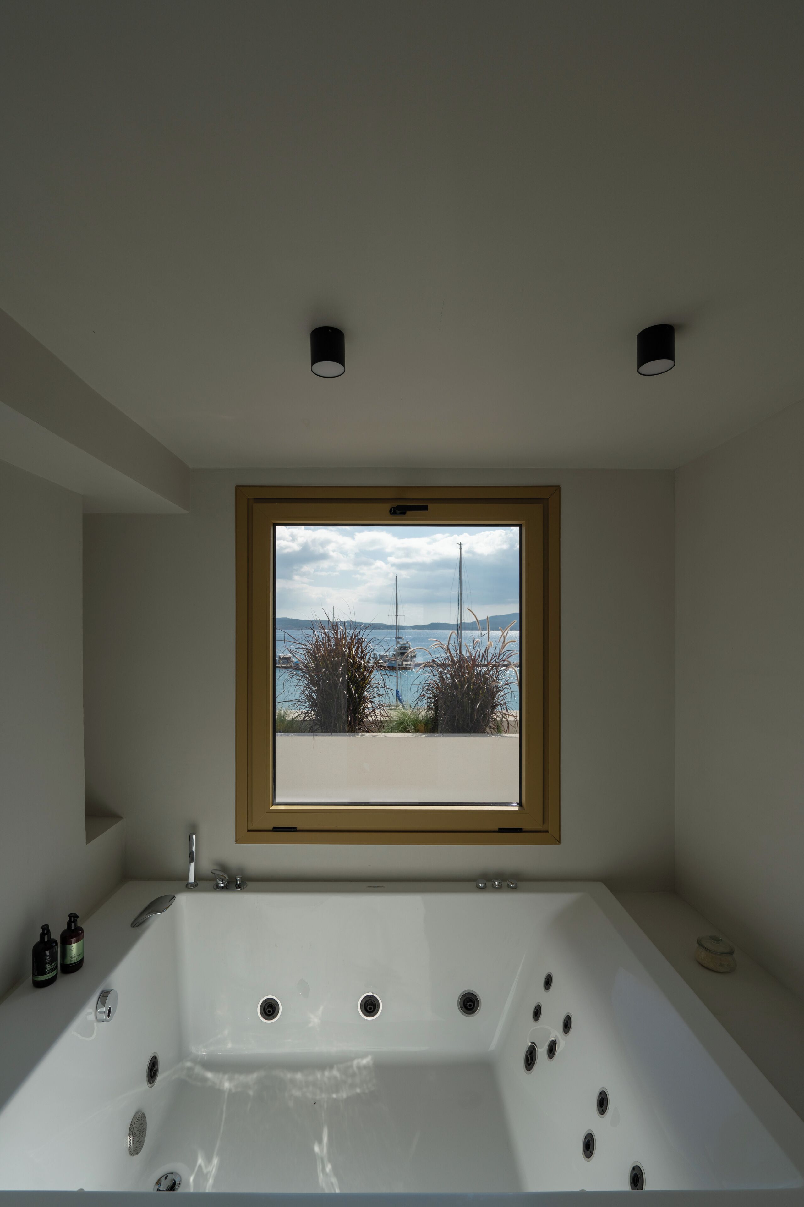 A modern bathroom with a large bathtub, a window with a view of the sea, and a small shelf with toiletries.
