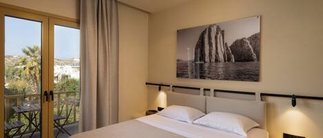 Habitación de hotel con cama, mesita de noche, lámpara de pared, ventana con cortinas y balcón con mesa y sillas.