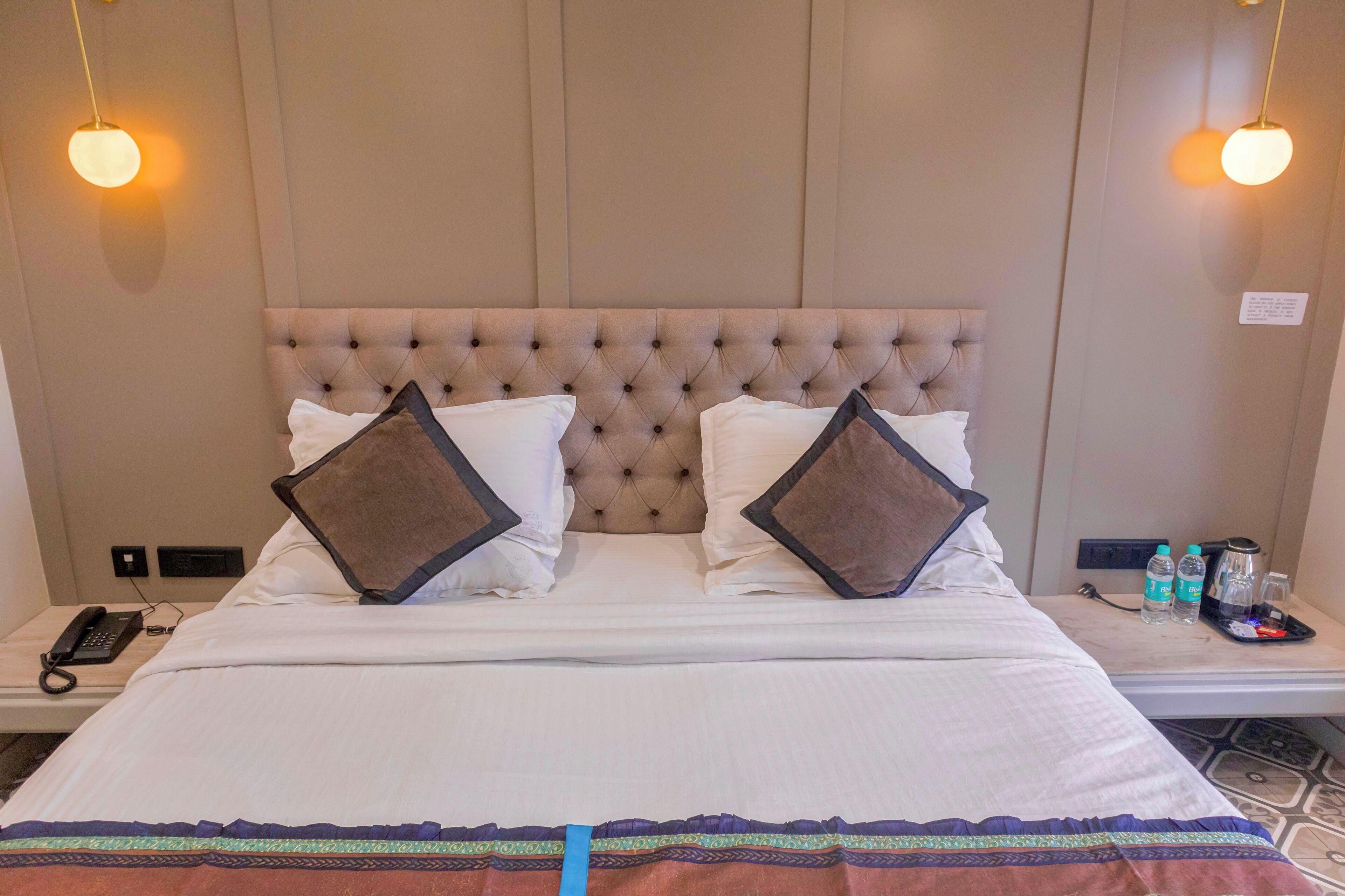 A hotel room with a tufted headboard, two pillows, a bedside table with a phone, water bottles, and a tray with a coffee pot.