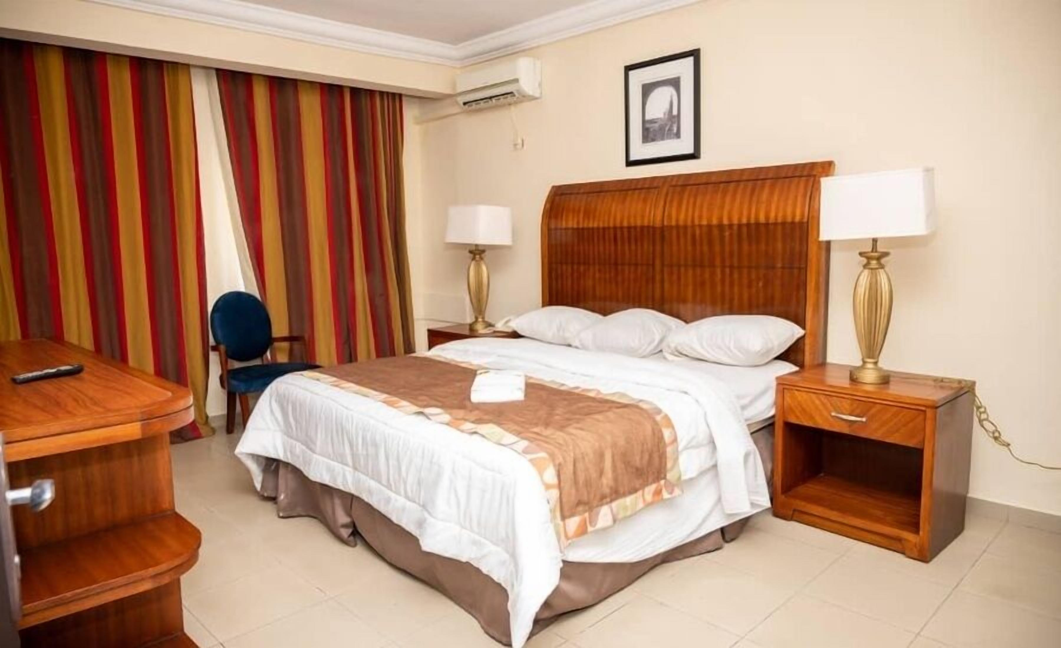A hotel room with a bed, two bedside tables with lamps, a desk, a chair, and a striped curtain.