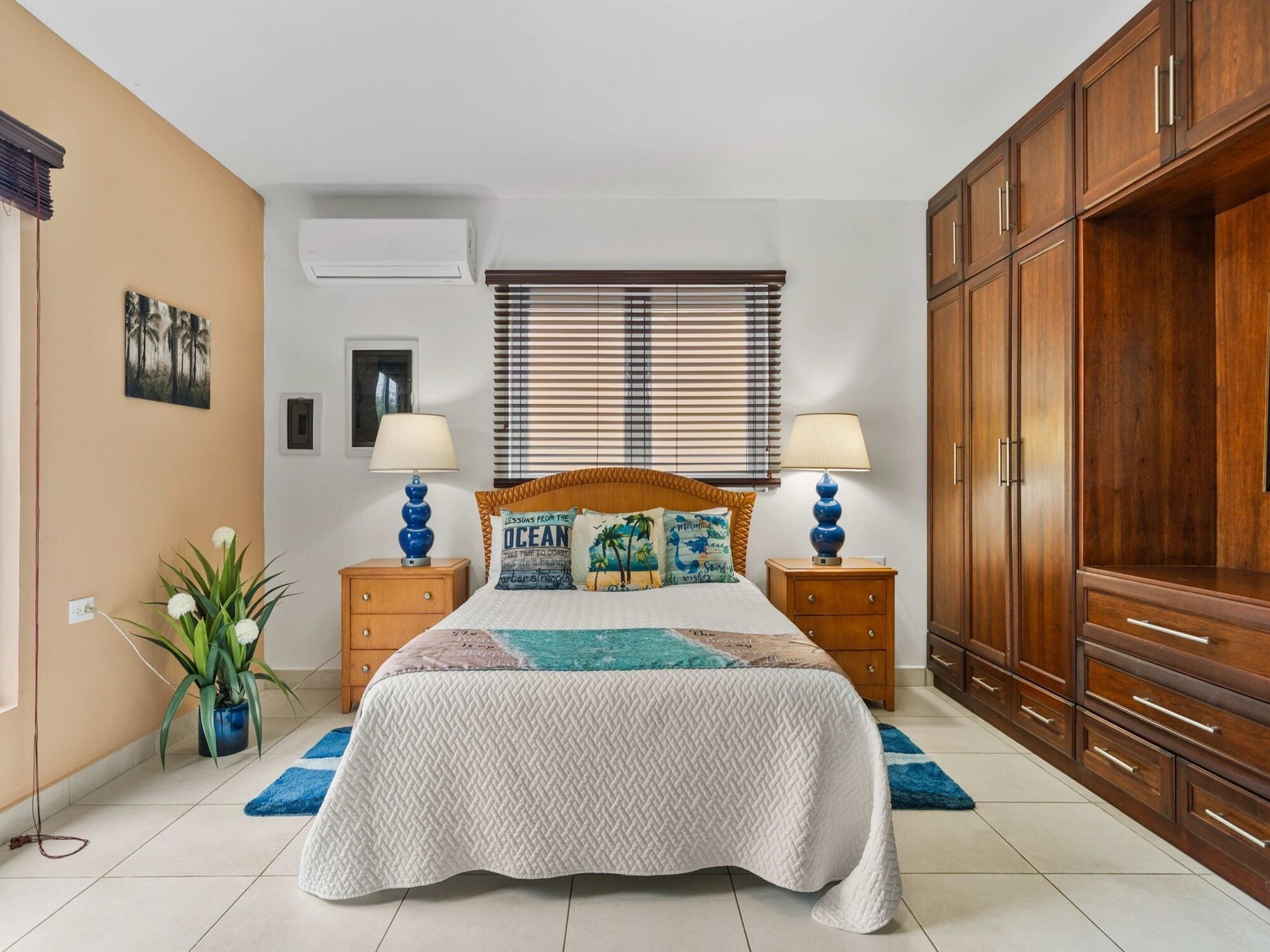 A bedroom with a bed, two bedside tables, a nightstand, a window with blinds, a wall-mounted air conditioner, a large wooden wardrobe, and a potted plant.