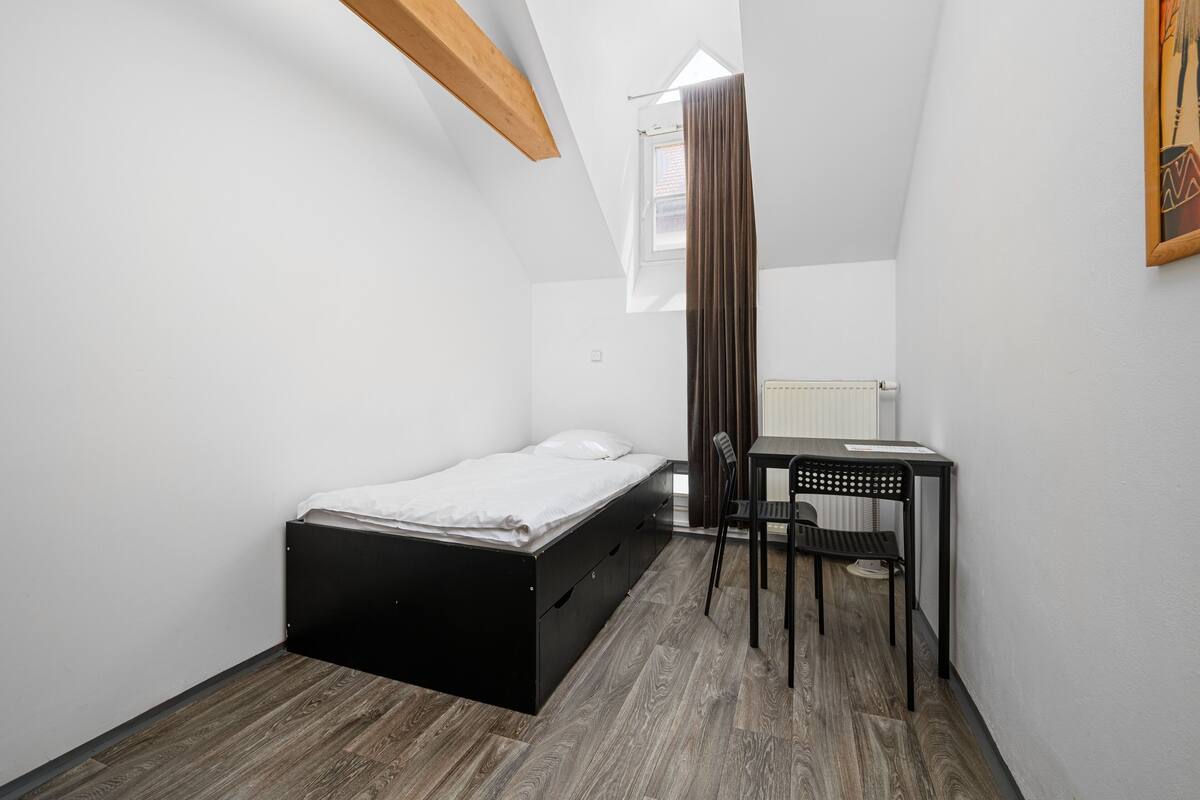 Una habitación pequeña y luminosa con una cama, un escritorio y una silla. Tiene una ventana con cortinas y un radiador en la pared.