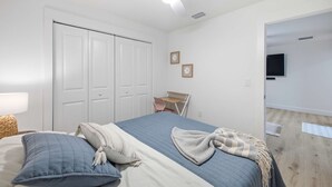 A bedroom with a bed, a ceiling fan, a wardrobe, a small table with a chair, and a wall-mounted TV.