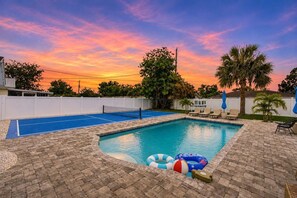 Een zwembad met een blauw terras, ligstoelen en een zonsondergang.