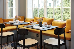 Un coin repas avec une table dressée pour deux personnes, un banc doté de coussins jaunes et une vue sur la verdure grâce à de grandes fenêtres.