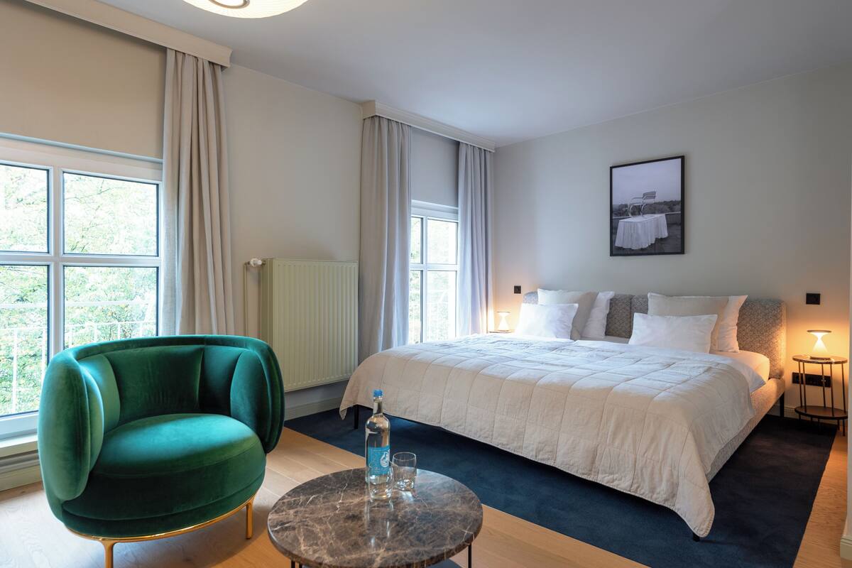 A hotel room with a large bed, a green armchair, a round table with a bottle and glass, and a view of greenery through the windows.