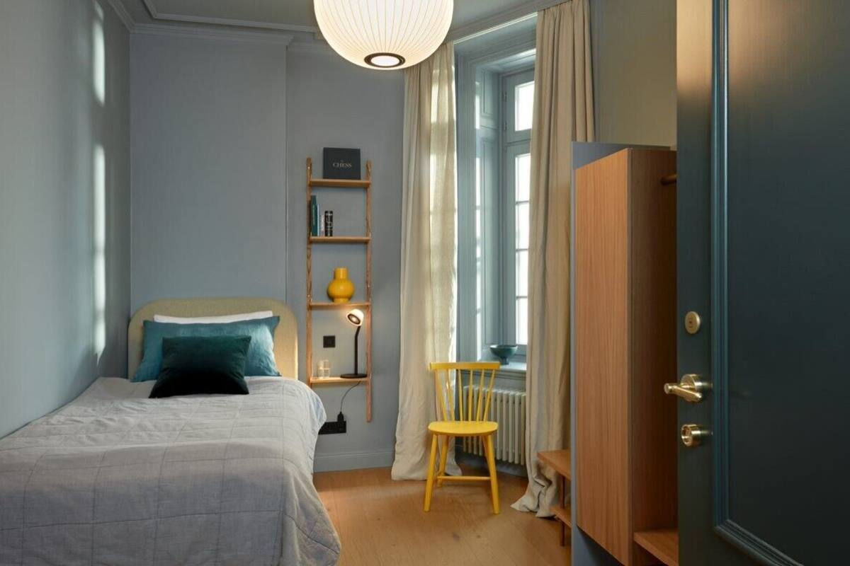 A bedroom with a bed, a wooden chair, a shelf with books and a lamp, and a wardrobe.