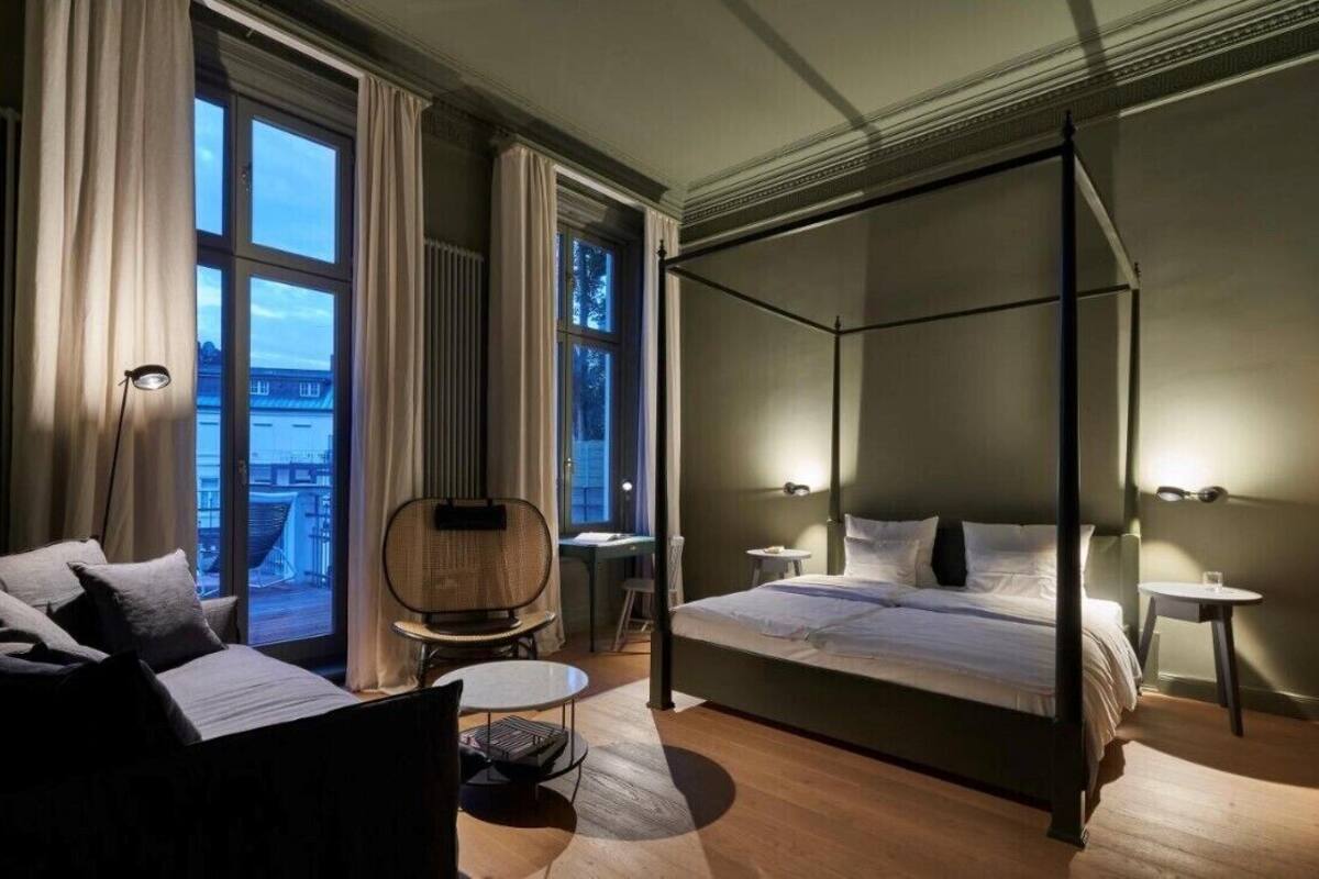 A modern bedroom with a four-poster bed, a sitting area with a chair and table, and large windows offering a view of the city.