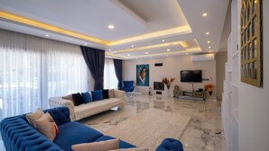 A modern living room with a sectional sofa, a white sofa, and a blue armchair. There is a flat-screen TV mounted on the wall, a coffee table, and a decorative vase with flowers. - Luxury Private Pool Villa in Fethiye6En-Suite Bedrooms Sleeps12  Sea&Nature View (Mugla)