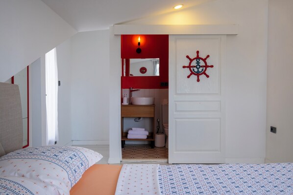 A bedroom with a bed, a nightstand, a red wall, a white door, and a small bathroom visible through the door.