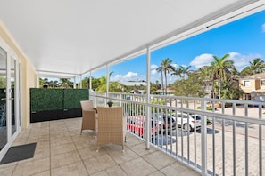 A covered balcony with wicker chairs, a green wall, and a view of parked cars and palm trees.