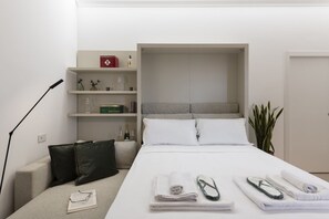Een hotelkamer met een bed, nachtkastje, lamp, plant en een plank met toiletartikelen.