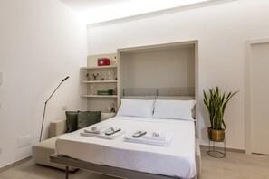 Ein modernes Hotelzimmer mit Bett, Nachttischen, einem Ablagefach mit Pflegeprodukten, einer Topfpflanze und einer Stehlampe.