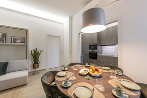 Ein moderner Essbereich mit einem runden Tisch für vier Personen, einer Couch, einem Blumentopf und einer Küche im Hintergrund.