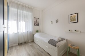 Une chambre à coucher avec un lit, une petite table, une étagère avec des plantes et une fenêtre ornée de rideaux légers.