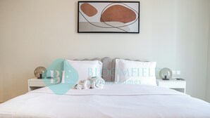 Een net opgemaakt bed met witte lakens en kussens, een nachtkastje met een lamp en een lijst met abstracte kunst aan de muur.