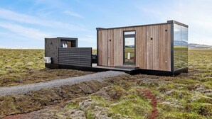 A modern cabin with a wooden exterior and a glass door, situated in a grassy field with mountains in the background. - Heathland Lodge (Selfoss)