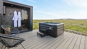 A modern outdoor deck with a hot tub, two lounge chairs, and a seating area with a view of the landscape. - Heathland Lodge (Selfoss)